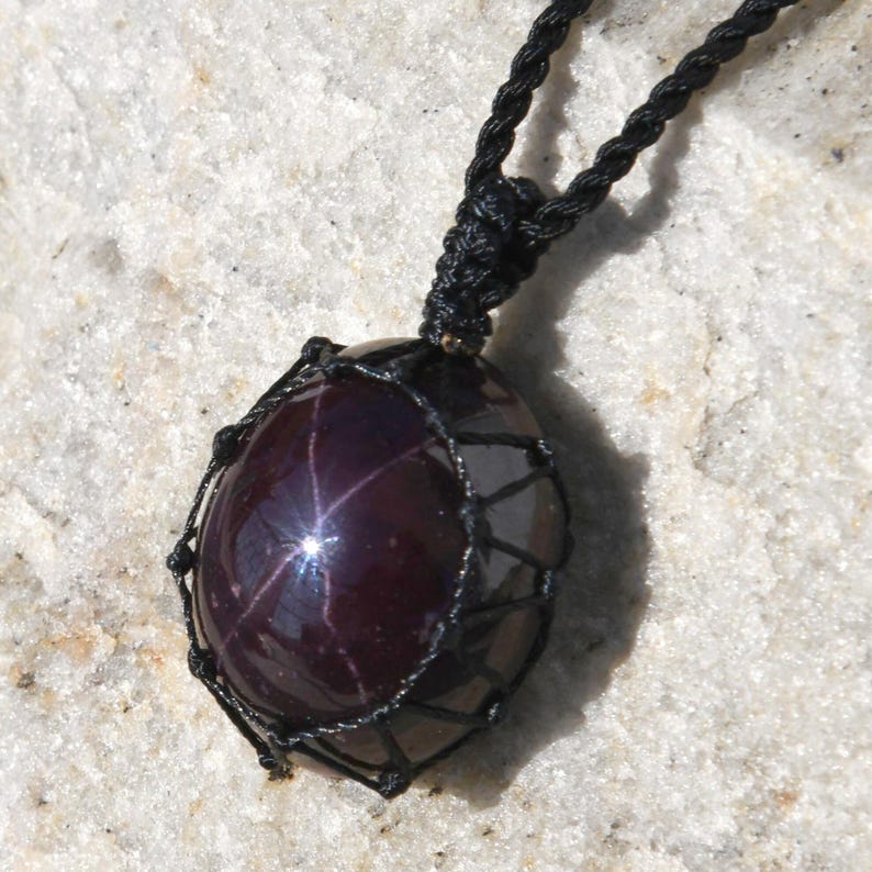 Close-up of a polished star garnet pendant in deep burgundy, wine-red, and dark plum tones, displayed on a light stone background. A distinct four-rayed star appears across the gemstone when illuminated, creating a bright reflective cross. The garnet has a smooth glossy finish with rich color depth and natural translucency. It is secured in a black macrame net and attached to a black adjustable cord. Associated with passion, vitality, grounding, courage, and emotional strength.