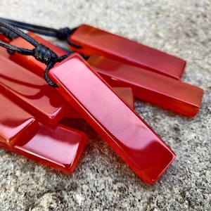Several polished, elongated, rectangular Carnelian crystal necklaces are displayed on a textured gray surface. The orange-red gemstones have a smooth, translucent texture. There are multiple pendants, each attached to a black cord. Carnelian is associated with courage, vitality, and motivation.