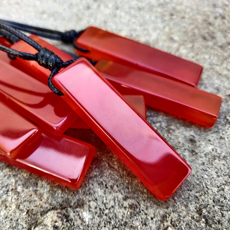 Several polished, elongated, rectangular Carnelian crystal necklaces are displayed on a textured gray surface. The orange-red gemstones have a smooth, translucent texture. There are multiple pendants, each attached to a black cord. Carnelian is associated with courage, vitality, and motivation.