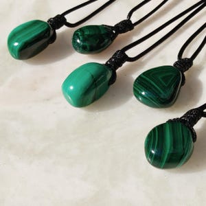 Close-up of 5 polished irregular Malachite in shades of vivid green and deep forest green, arranged on a light stone surface. The green opaque gemstones have smooth glossy surfaces with distinct banded patterns, concentric lines, and varied formations, including rounded and triangular shapes with layered green stripes and darker swirls. It is attached to a black cord with a top knot and adjustable knot closure. Malachite is associated with transformation, protection, and emotional healing