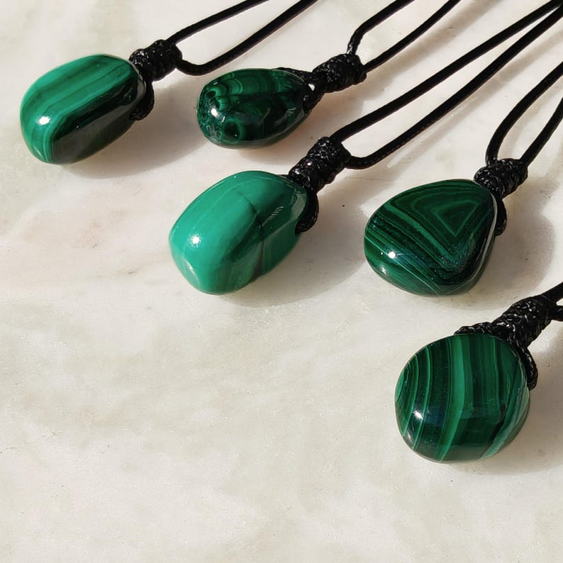 Close-up of 5 polished irregular Malachite in shades of vivid green and deep forest green, arranged on a light stone surface. The green opaque gemstones have smooth glossy surfaces with distinct banded patterns, concentric lines, and varied formations, including rounded and triangular shapes with layered green stripes and darker swirls. It is attached to a black cord with a top knot and adjustable knot closure. Malachite is associated with transformation, protection, and emotional healing