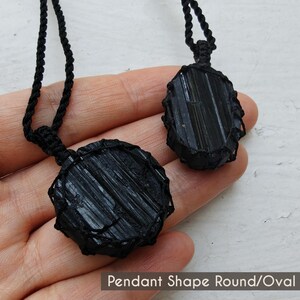 Close-up of 2 raw Black Tourmaline in deep black shades, displayed on a hand against a white background. The Black Tourmaline dark gemstone shows strong parallel vertical striations, rough uneven edges, and a matte to slightly reflective texture with natural ridges and minor surface fractures. One piece is round and the other oval, both wrapped in a black macrame net and attached to a black cord with an adjustable knot closure. The image includes the text “Pendant Shape Round/Oval”.