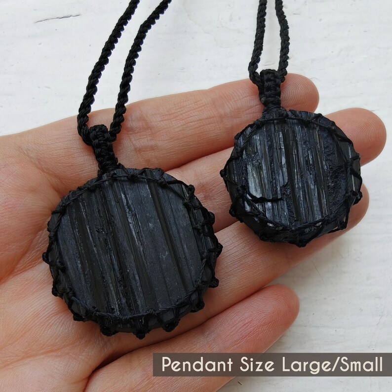 Close-up of 2 raw round Black Tourmaline in deep black shades, displayed on a hand against a white background. The Black Tourmaline dark gemstone has strong parallel vertical striations, rough uneven edges, and a matte to slightly reflective texture with natural ridges and minor surface fractures. Each piece is wrapped in a black macrame net and attached to a black cord with an adjustable knot closure. The image includes the text “Pendant Size Large/Small”.