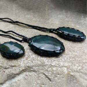 Three Bloodstone pendant necklaces displayed against a textured natural stone attached to adjustable twisted black cords with a sliding knot closure. Each polished oval gemstone features deep forest-green tones with subtle natural red mineral inclusions, making every pendant unique. Bloodstone is associated with Courage, Resilience, Grounding, Vitality, and Inner Strength.
