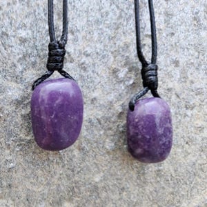 Two smooth, polished, rounded Lepidolite necklace pendants are displayed on a textured gray concrete surface. The purple gemstones vary slightly in color intensity and show some lighter patches and subtle striations. Each pendant is attached to a black cord. Lepidolite is associated with self-love, anti-anxiety, stress relief, transformation, emotional healing, and negativity release.