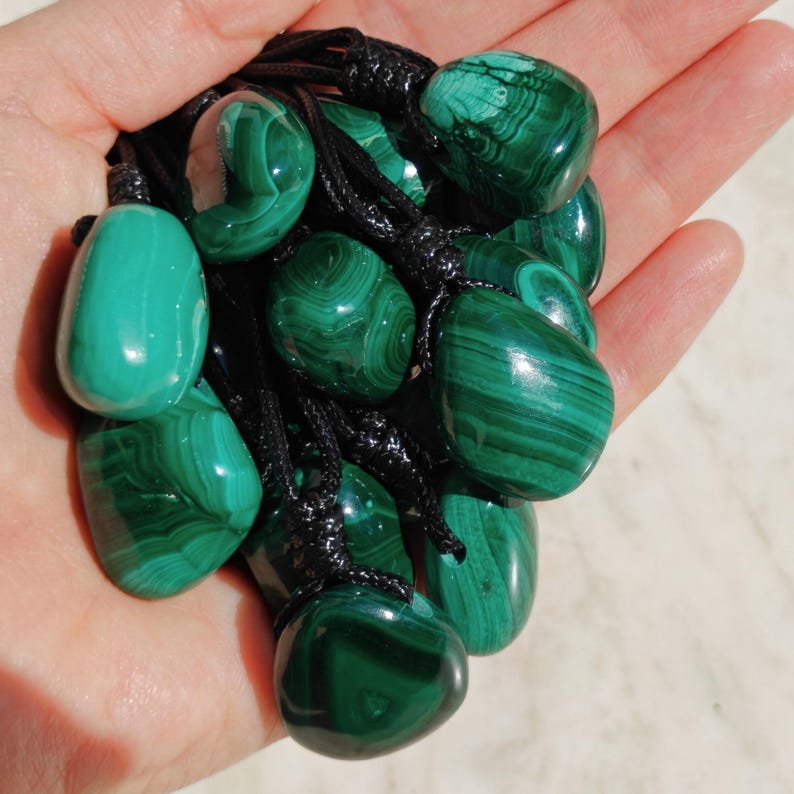 Close-up of multiple polished irregular Malachite in shades of vivid green and deep forest green, held in a hand against a light background. The green opaque gemstones have smooth glossy surfaces with distinct banded patterns, concentric rings, and flowing lines, with varied shapes and layered green formations visible across the stones. It is attached to a black cord with a top knot and adjustable knot closure. Malachite is associated with transformation, protection, and emotional healing.