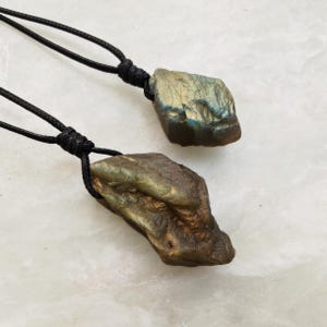 Close-up of two raw irregular Labradorite in shades of yellow and golden brown, resting on a light stone surface. The Labradorite gemstones have a natural rough texture with strong iridescent flashes, visible striations, and layered color play with reflective highlights. They are secured with a top knot and attached to black cords with adjustable knot closure. Labradorite is associated with transformation, protection, intuition, and inner strength.