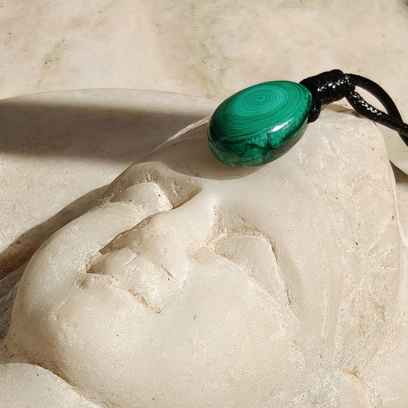 Close-up of a polished oval Malachite in shades of vivid green and deep forest green, resting on a carved light stone surface. The green opaque gemstone has a smooth glossy surface with distinct concentric banding, circular eye-like formations, and layered green gradients, with light reflections highlighting its rounded shape. It is attached to a black cord with a top knot and adjustable knot closure. Malachite is associated with transformation, protection, and emotional healing.
