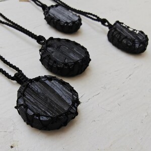 Close-up of 4 raw oval and round Black Tourmaline in deep black shades, resting on a white wood surface. The Black Tourmaline dark gemstone has strong parallel vertical striations, rough uneven edges, and a matte to slightly reflective texture with natural ridges and occasional lighter inclusions and surface variations. Each piece is wrapped in a black macrame net and attached to a black cord with an adjustable knot closure. Black Tourmaline is associated with grounding and protection.