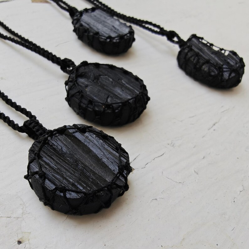 Close-up of 4 raw oval and round Black Tourmaline in deep black shades, resting on a white wood surface. The Black Tourmaline dark gemstone has strong parallel vertical striations, rough uneven edges, and a matte to slightly reflective texture with natural ridges and occasional lighter inclusions and surface variations. Each piece is wrapped in a black macrame net and attached to a black cord with an adjustable knot closure. Black Tourmaline is associated with grounding and protection.