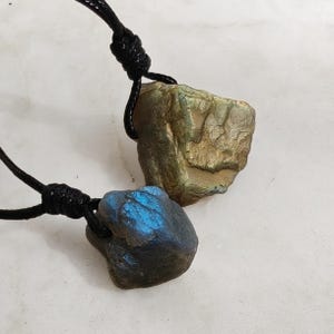 Close-up of two raw irregular Labradorite in shades of blue, teal, and golden brown, resting on a light stone surface. The Labradorite gemstones have a natural rough texture with strong iridescent flashes, visible striations, and layered color play with reflective highlights. They are secured with a top knot and attached to black cords with adjustable knot closure. Labradorite is associated with transformation, protection, intuition, and inner strength.