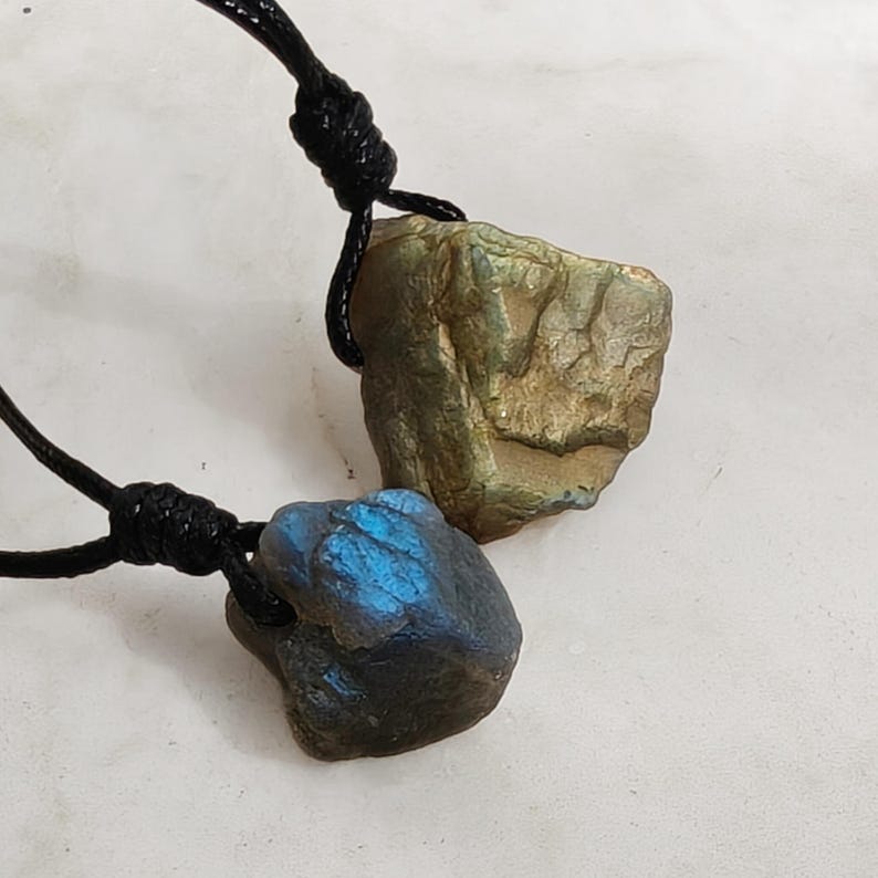 Close-up of two raw irregular Labradorite in shades of blue, teal, and golden brown, resting on a light stone surface. The Labradorite gemstones have a natural rough texture with strong iridescent flashes, visible striations, and layered color play with reflective highlights. They are secured with a top knot and attached to black cords with adjustable knot closure. Labradorite is associated with transformation, protection, intuition, and inner strength.