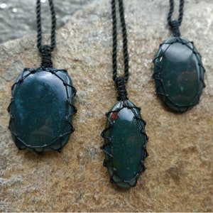 Three Bloodstone pendant necklaces displayed hanging from adjustable twisted black cords with a sliding knot closure against a textured natural stone wall. Each polished oval gemstone features deep forest-green tones with subtle natural red mineral inclusions, making every pendant unique. Bloodstone is associated with Courage, Resilience, Grounding, Vitality, and Inner Strength.