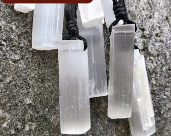 Puede incluir: Colgantes de cristal de selenita en cordones negros. Los colgantes son rectangulares y translúcidos, con estrías internas visibles. El fondo es una superficie de piedra gris rugosa. El texto "$25 CLEARANCE" está en un rectángulo marrón.