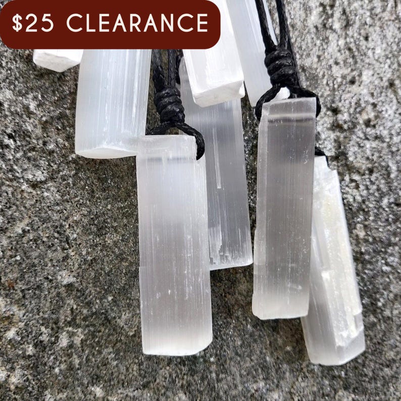Several raw, rectangular selenite pendants hang from black cord necklaces, clustered together on a textured gray stone surface. The translucent white crystals have a fibrous, slightly uneven texture and catch the light. These selenite pendants are associated with clarity and purification.