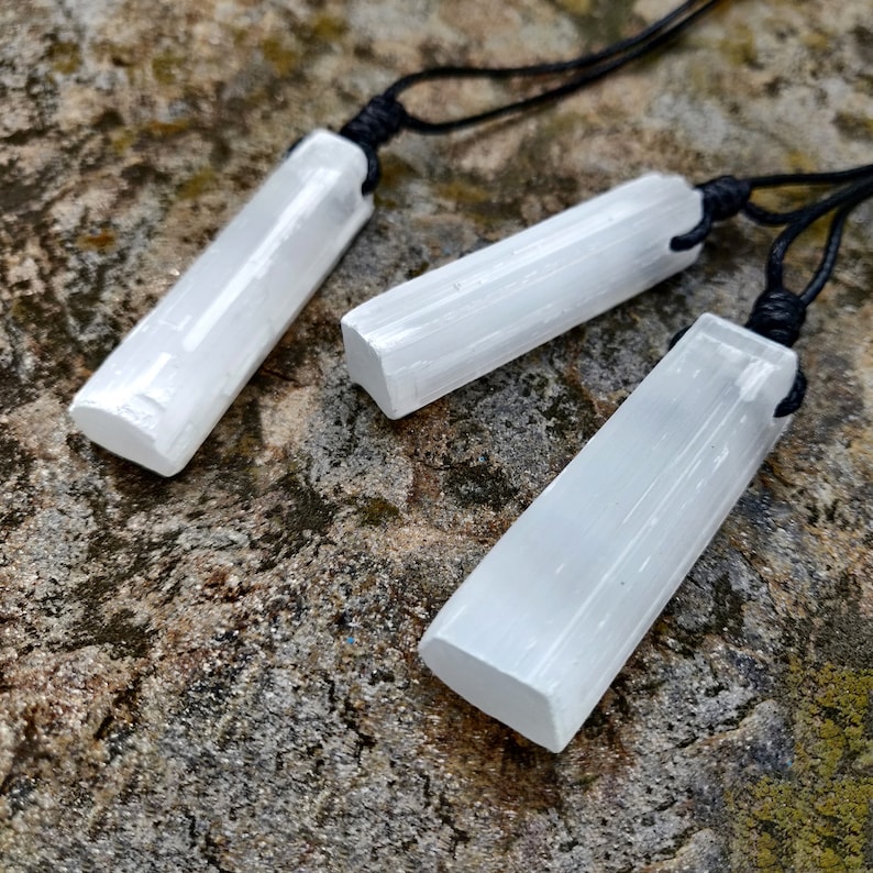 Three raw, rectangular selenite pendants hang from black cord necklaces, clustered together on a textured gray stone surface. The translucent white crystals have a fibrous, slightly uneven texture and catch the light. These selenite pendants are associated with clarity and purification.