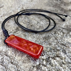 May include: A rectangular, reddish-orange pendant necklace with a glossy finish. The pendant is suspended from a black cord, knotted at the top and adjustable. The necklace rests on a textured, gray stone surface.