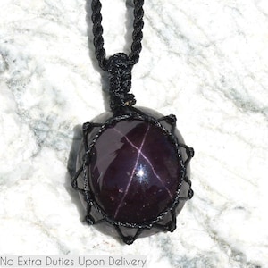 A single, smooth, polished, oval Almandine Star Garnet necklace pendant is displayed on a textured white marble surface. The deep purple-red gemstone exhibits a distinct four-rayed star (asterism) in its center when light reflects off it. Almandine Star Garnet is associated with inner strength, protection, vitality, and regeneration. It is believed to inspire love and devotion, and to balance energy. It is encased in a black macrame net that forms the necklace cord.