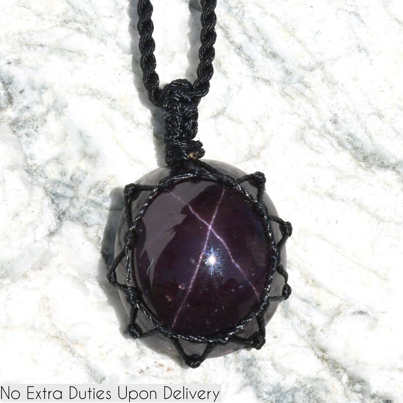 A single, smooth, polished, oval Almandine Star Garnet necklace pendant is displayed on a textured white marble surface. The deep purple-red gemstone exhibits a distinct four-rayed star (asterism) in its center when light reflects off it. Almandine Star Garnet is associated with inner strength, protection, vitality, and regeneration. It is believed to inspire love and devotion, and to balance energy. It is encased in a black macrame net that forms the necklace cord.