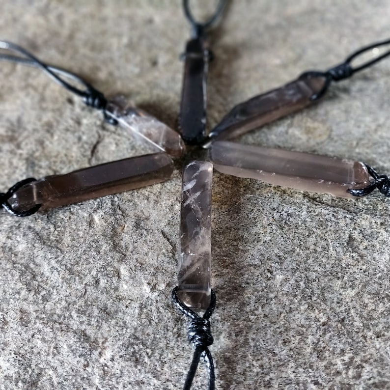Six raw smoky quartz necklaces with rectangular stones in dark brown translucent hues, rough-textured with irregular edges, each hung on a knotted black cord, placed on a light brown stone surface with a natural grainy texture. Smokey Quartz is associated with grounding, protection, and emotional calmness.