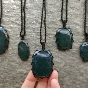 Several Bloodstone pendant necklaces displayed hanging from adjustable twisted black cords with a sliding knot closure against a textured natural stone wall. Each polished oval gemstone features deep forest-green tones with subtle natural red mineral inclusions, making every pendant unique. Bloodstone is associated with Courage, Resilience, Grounding, Vitality, and Inner Strength.