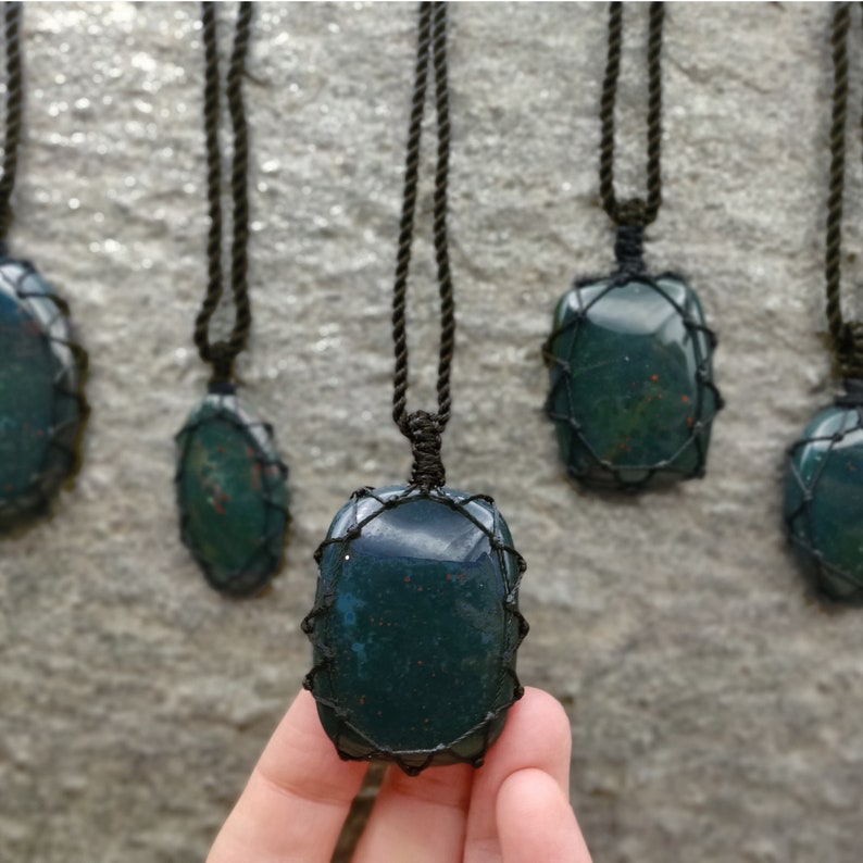Several Bloodstone pendant necklaces displayed hanging from adjustable twisted black cords with a sliding knot closure against a textured natural stone wall. Each polished oval gemstone features deep forest-green tones with subtle natural red mineral inclusions, making every pendant unique. Bloodstone is associated with Courage, Resilience, Grounding, Vitality, and Inner Strength.