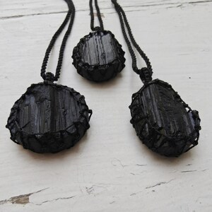 Close-up of 3 raw oval and round Black Tourmaline in deep black shades, resting on a white wood surface. The Black Tourmaline dark gemstone has strong parallel vertical striations, rough uneven edges, and a matte to slightly reflective texture with natural ridges and occasional lighter inclusions and surface variations. Each piece is wrapped in a black macrame net and attached to a black cord with an adjustable knot closure. Black Tourmaline is associated with grounding and protection.