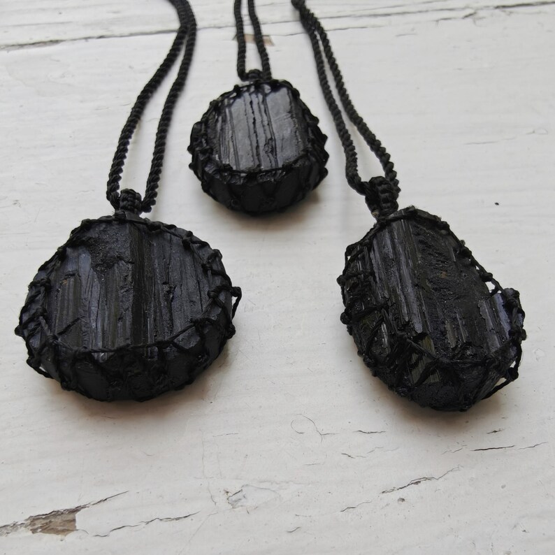 Close-up of 3 raw oval and round Black Tourmaline in deep black shades, resting on a white wood surface. The Black Tourmaline dark gemstone has strong parallel vertical striations, rough uneven edges, and a matte to slightly reflective texture with natural ridges and occasional lighter inclusions and surface variations. Each piece is wrapped in a black macrame net and attached to a black cord with an adjustable knot closure. Black Tourmaline is associated with grounding and protection.