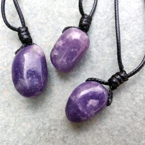 Three smooth, polished, rounded Lepidolite necklace pendants are displayed on a textured gray concrete surface. The purple gemstones vary slightly in color intensity and show some lighter patches and subtle striations. Each pendant is attached to a black cord. Lepidolite is associated with self-love, anti-anxiety, stress relief, transformation, emotional healing, and negativity release.