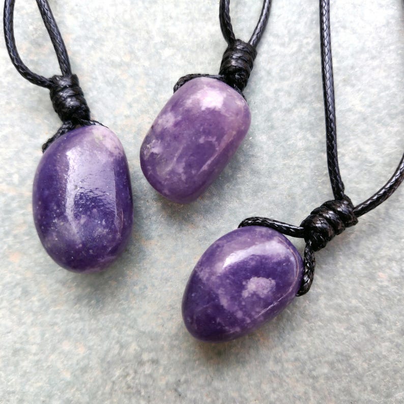 Three smooth, polished, rounded Lepidolite necklace pendants are displayed on a textured gray concrete surface. The purple gemstones vary slightly in color intensity and show some lighter patches and subtle striations. Each pendant is attached to a black cord. Lepidolite is associated with self-love, anti-anxiety, stress relief, transformation, emotional healing, and negativity release.