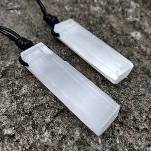 Two raw, rectangular selenite pendants hang from black cord necklaces, clustered together on a textured gray stone surface. The translucent white crystals have a fibrous, slightly uneven texture and catch the light. These selenite pendants are associated with clarity and purification.
