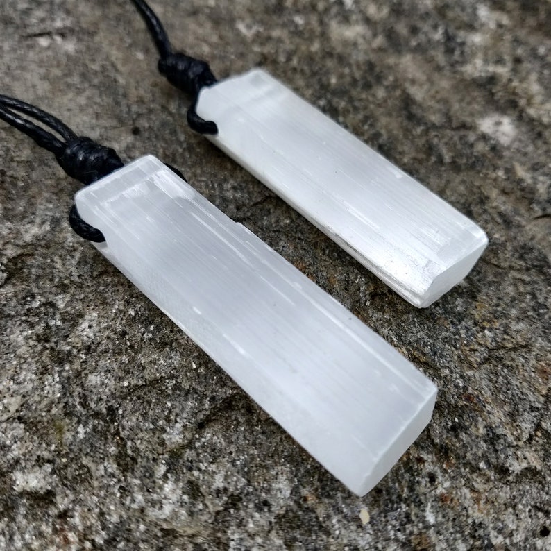 Two raw, rectangular selenite pendants hang from black cord necklaces, clustered together on a textured gray stone surface. The translucent white crystals have a fibrous, slightly uneven texture and catch the light. These selenite pendants are associated with clarity and purification.