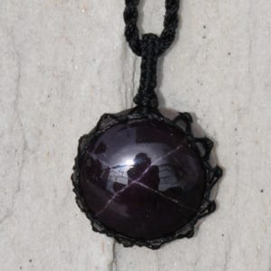A single, smooth, polished, oval Star Garnet necklace pendant is displayed on a textured light rock surface. The dark red to reddish-purple gemstone exhibits a distinct four-rayed star (asterism) in its center when light reflects off it. It is encased in a black macrame net that forms the necklace cord. Star Garnet is associated with inner strength, protection, vitality, and regeneration.