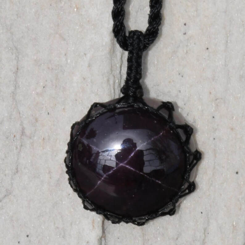 A single, smooth, polished, oval Star Garnet necklace pendant is displayed on a textured light rock surface. The dark red to reddish-purple gemstone exhibits a distinct four-rayed star (asterism) in its center when light reflects off it. It is encased in a black macrame net that forms the necklace cord. Star Garnet is associated with inner strength, protection, vitality, and regeneration.