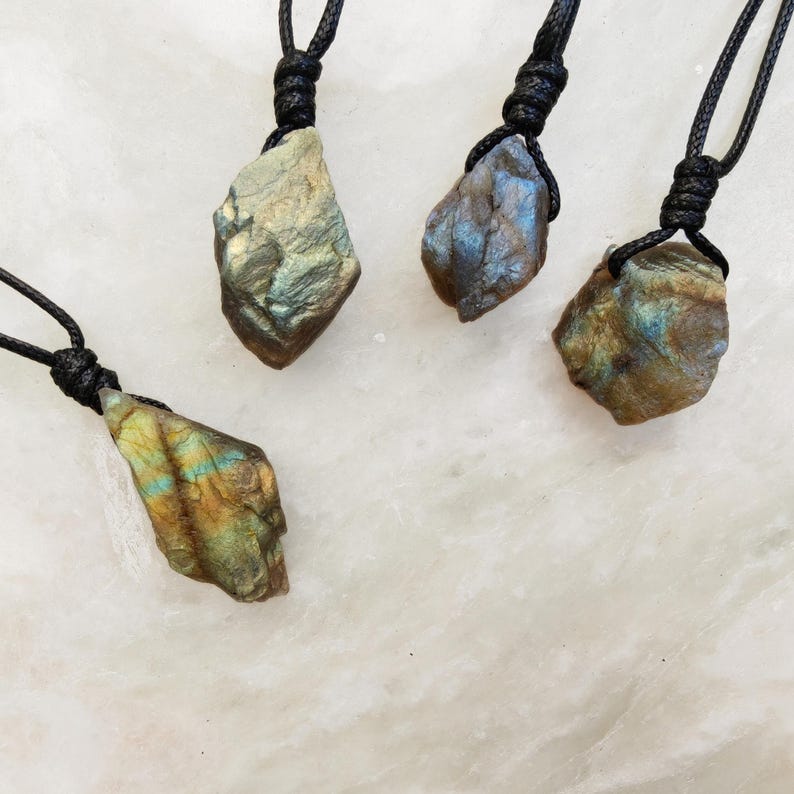 Close-up of 4 raw irregular Labradorite in shades of blue, teal, and golden brown, resting on a light stone surface. The Labradorite gemstones have a natural rough texture with strong iridescent flashes, visible striations, and layered color play with reflective highlights. They are secured with a top knot and attached to black cords with adjustable knot closure. Labradorite is associated with transformation, protection, intuition, and inner strength.