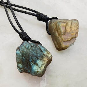 Close-up of two raw irregular Labradorite in shades of blue, teal, and golden brown, resting on a light stone surface. The Labradorite gemstones have a natural rough texture with strong iridescent flashes, visible striations, and layered color play with reflective highlights. They are secured with a top knot and attached to black cords with adjustable knot closure. Labradorite is associated with transformation, protection, intuition, and inner strength.
