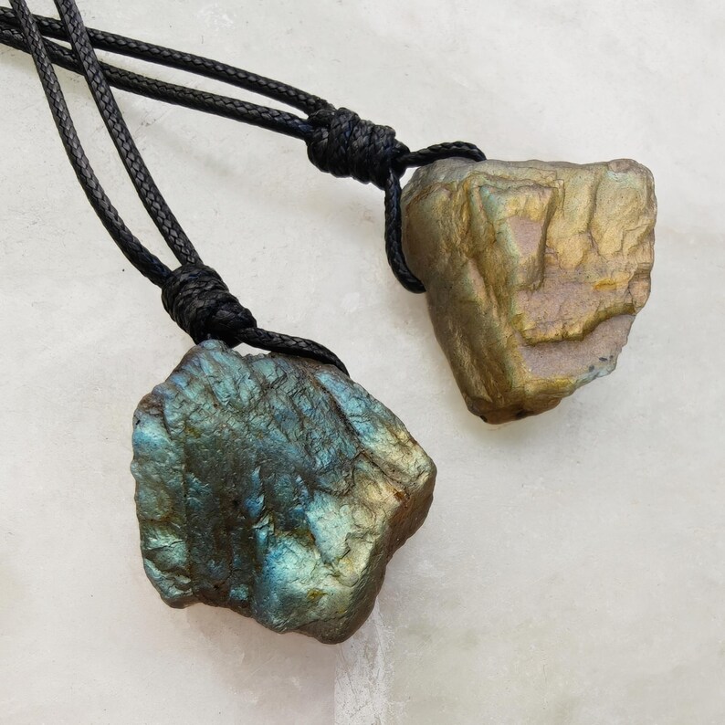 Close-up of two raw irregular Labradorite in shades of blue, teal, and golden brown, resting on a light stone surface. The Labradorite gemstones have a natural rough texture with strong iridescent flashes, visible striations, and layered color play with reflective highlights. They are secured with a top knot and attached to black cords with adjustable knot closure. Labradorite is associated with transformation, protection, intuition, and inner strength.