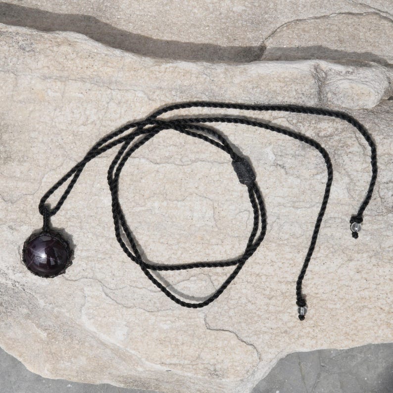 A single, smooth, polished, oval Star Garnet necklace pendant is displayed on a textured light rock surface. The dark red to reddish-purple gemstone exhibits a distinct four-rayed star (asterism) in its center when light reflects off it. It is encased in a black macrame net that forms the necklace cord. Star Garnet is associated with inner strength, protection, vitality, and regeneration.