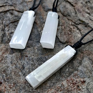 Three raw, rectangular selenite pendants hang from black cord necklaces, clustered together on a textured gray stone surface. The translucent white crystals have a fibrous, slightly uneven texture and catch the light. These selenite pendants are associated with clarity and purification.