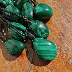 Close-up of multiple polished irregular Malachite in shades of vivid green and deep forest green, clustered together against a textured wood background. The green opaque gemstones have smooth glossy surfaces with distinct banded patterns, flowing concentric lines, and eye-like formations with darker green swirls. It is attached to a black cord with a top knot and adjustable knot closure. Malachite is associated with transformation, protection, and emotional healing.