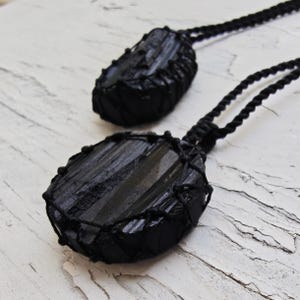 Close-up of 2 raw oval and round Black Tourmaline in deep black shades, resting on a white wood surface. The Black Tourmaline dark gemstone has strong parallel vertical striations, rough uneven edges, and a matte to slightly reflective texture with natural ridges and occasional lighter inclusions and surface variations. Each piece is wrapped in a black macrame net and attached to a black cord with an adjustable knot closure. Black Tourmaline is associated with grounding and protection.