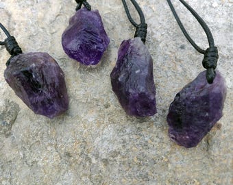 May include: Four raw amethyst crystal pendants on black cords. The amethyst stones are a deep purple color and have a rough, natural texture. The cords are knotted at the top, creating a simple, earthy design. The background is a light gray stone surface.
