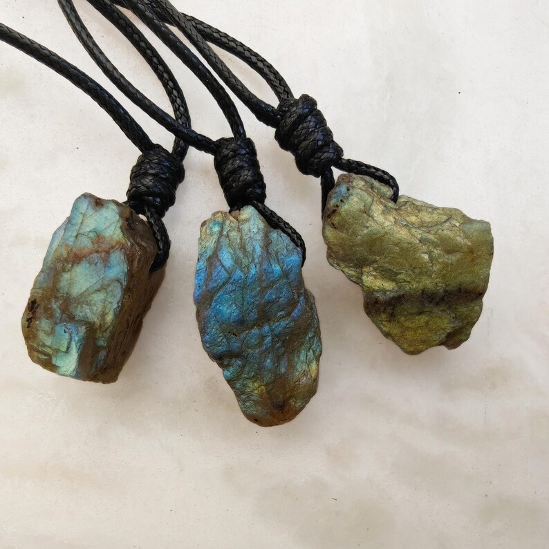 Close-up of 3 raw irregular Labradorite in shades of blue, teal, and golden brown, resting on a light stone surface. The Labradorite gemstones have a natural rough texture with strong iridescent flashes, visible striations, and layered color play with reflective highlights. They are secured with a top knot and attached to black cords with adjustable knot closure. Labradorite is associated with transformation, protection, intuition, and inner strength.