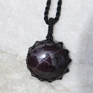 A single, smooth, polished, oval Star Garnet necklace pendant is displayed on a textured light rock surface. The dark red to reddish-purple gemstone exhibits a distinct four-rayed star (asterism) in its center when light reflects off it. It is encased in a black macrame net that forms the necklace cord. Star Garnet is associated with inner strength, protection, vitality, and regeneration.