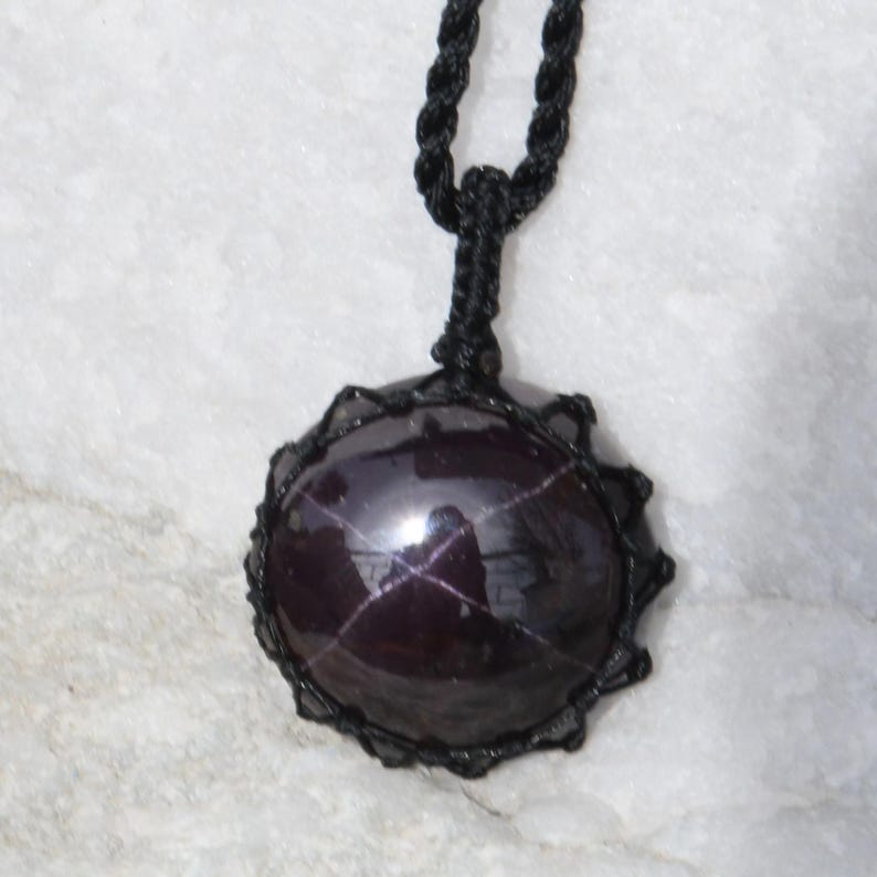 A single, smooth, polished, oval Star Garnet necklace pendant is displayed on a textured light rock surface. The dark red to reddish-purple gemstone exhibits a distinct four-rayed star (asterism) in its center when light reflects off it. It is encased in a black macrame net that forms the necklace cord. Star Garnet is associated with inner strength, protection, vitality, and regeneration.