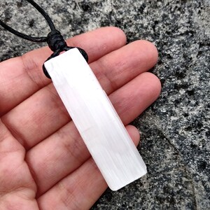 One raw, rectangular selenite pendant hang from black cord necklaces, rest on hand palm over a textured gray stone surface background. The translucent white crystal have a fibrous, slightly uneven texture and catch the light. These selenite pendants are associated with clarity and purification.