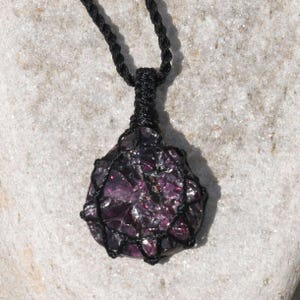 A single, raw, dtop shaped raw Garnet necklace pendant is displayed on a textured white rock surface. The dark reddish-purple gemstone has a rough, unpolished texture. It is encased in a black macrame net that forms the necklace cord. Garnet is associated with passion, energy, protection, and healing.