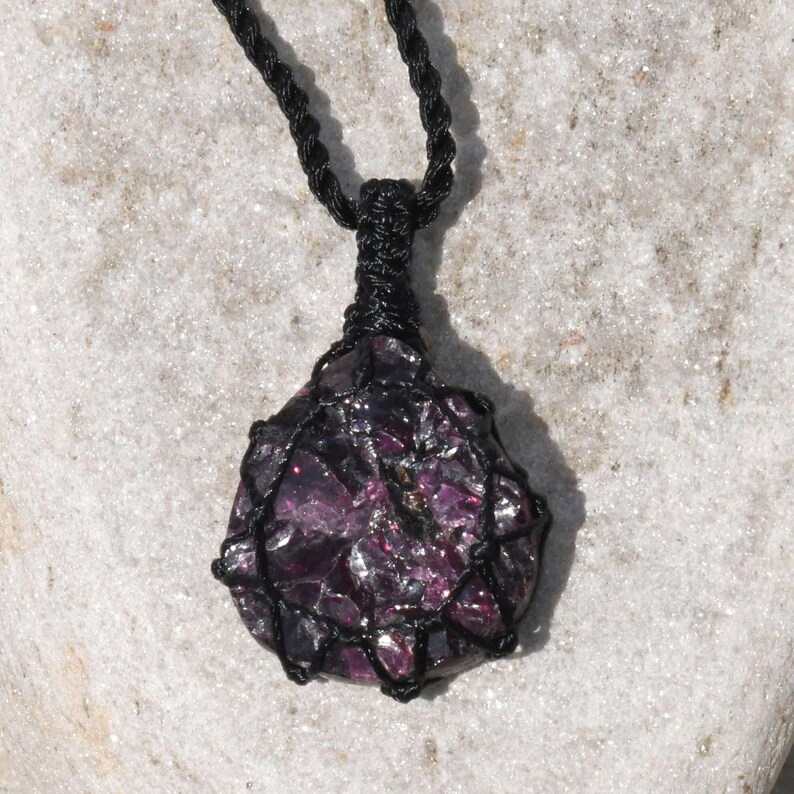 A single, raw, dtop shaped raw Garnet necklace pendant is displayed on a textured white rock surface. The dark reddish-purple gemstone has a rough, unpolished texture. It is encased in a black macrame net that forms the necklace cord. Garnet is associated with passion, energy, protection, and healing.