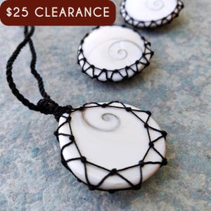 May include: A white pendant necklace with a spiral design, encased in a black woven net. The necklace is displayed with two matching earrings. The background is a textured blue-green surface.