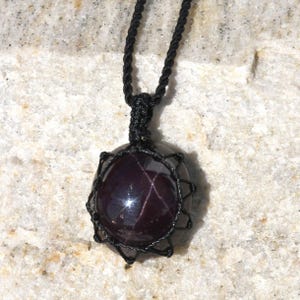 Close-up of a polished star garnet pendant in deep burgundy, wine-red, and dark plum tones, displayed on a light stone background. A distinct four-rayed star appears across the gemstone when illuminated, creating a bright reflective cross. The garnet has a smooth glossy finish with rich color depth and natural translucency. It is secured in a black macrame net and attached to a black adjustable cord. Associated with passion, vitality, grounding, courage, and emotional strength.